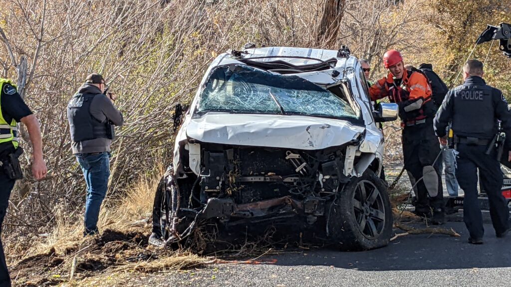 White damaged car with a broken windshield and a firefighter in orange standing next to it